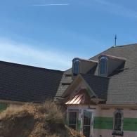 A new house under construction featuring gray shingles, arched dormer windows, and a copper roof detail above a doorway.