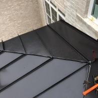 A high-angle view of a newly installed dark gray standing seam metal roof section with black metal joinery.