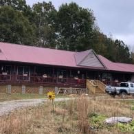 A one-story log cabin with a red metal roof and a wrap-around wooden porch sits on a grassy lot with a pickup truck nearby.
