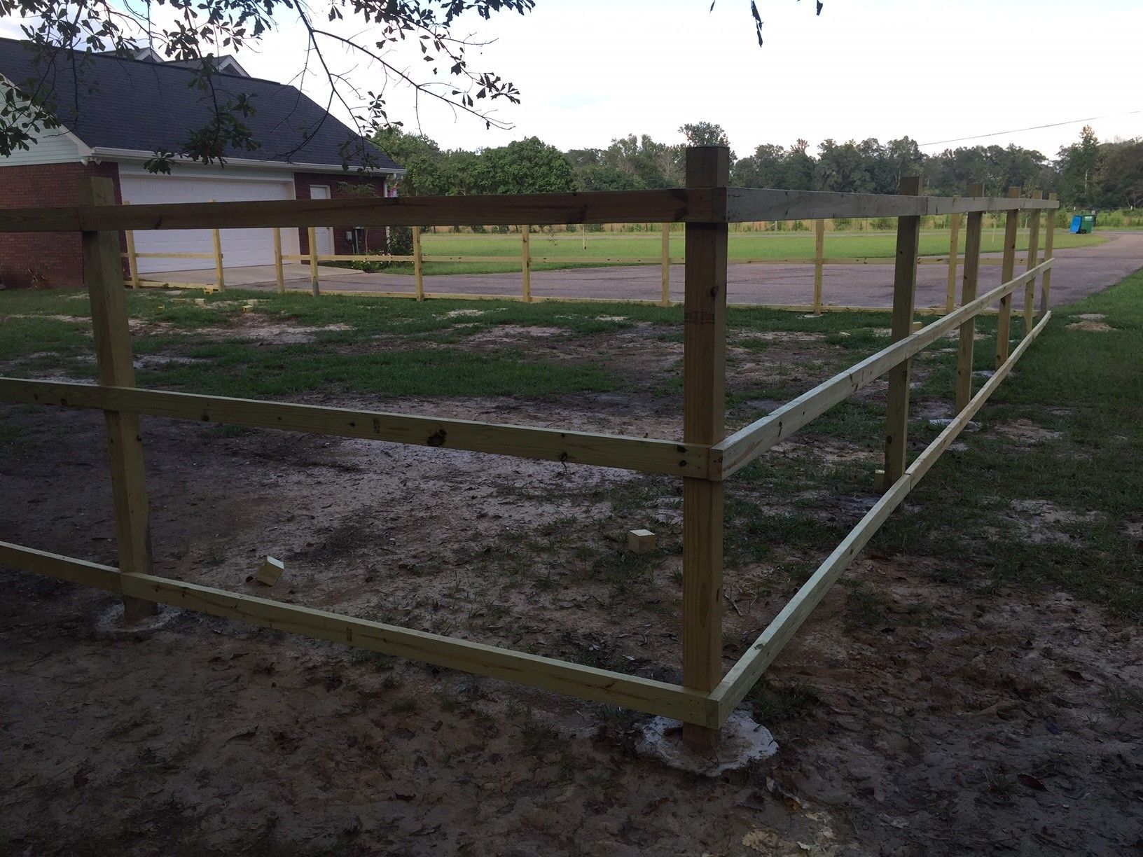 Wooden-and-metal fence enclosing a muddy paddock beside a grassy field and rural road.
