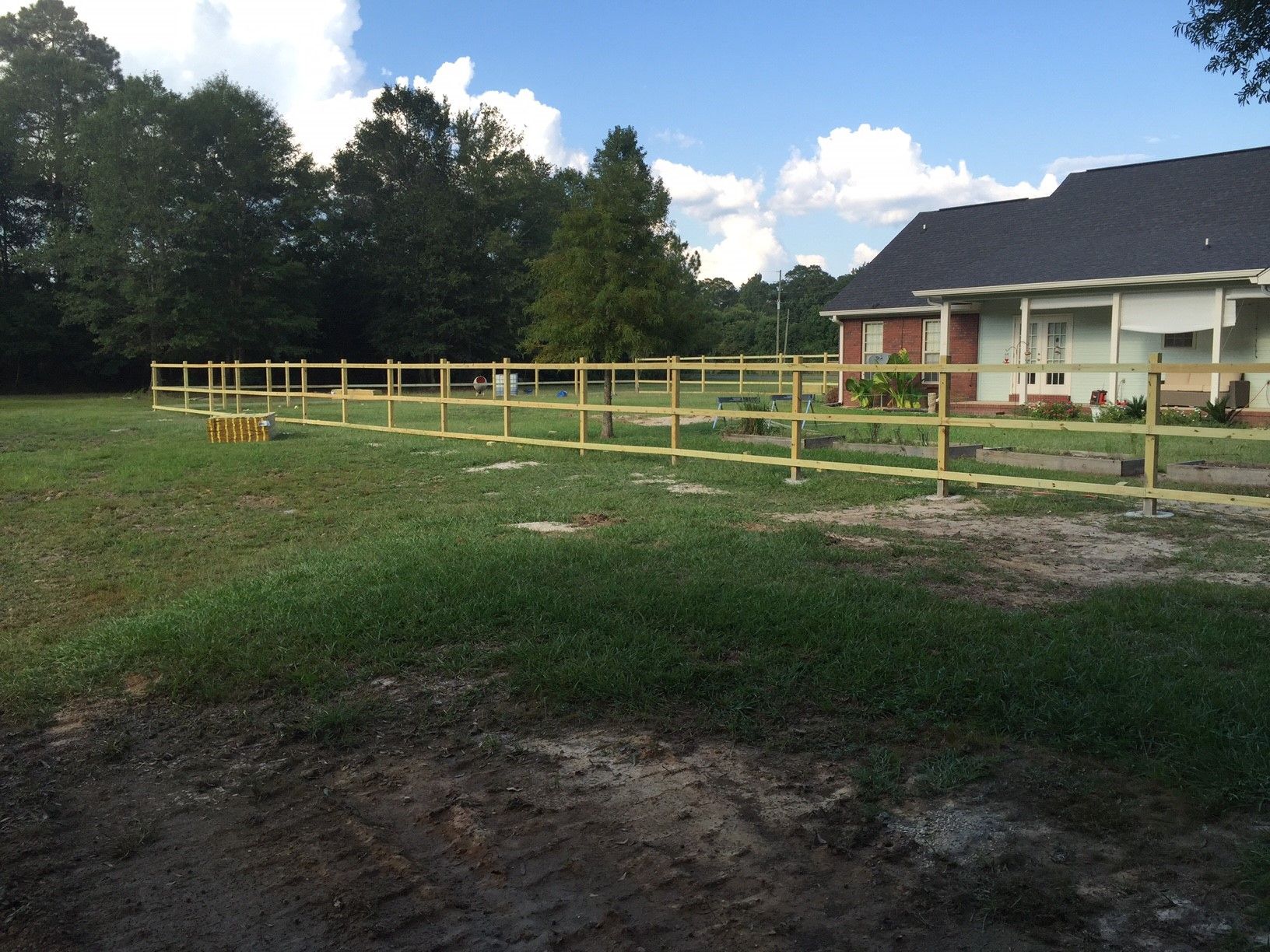 Rural yard with a wooden fence, grassy field, and a white house under a blue sky
