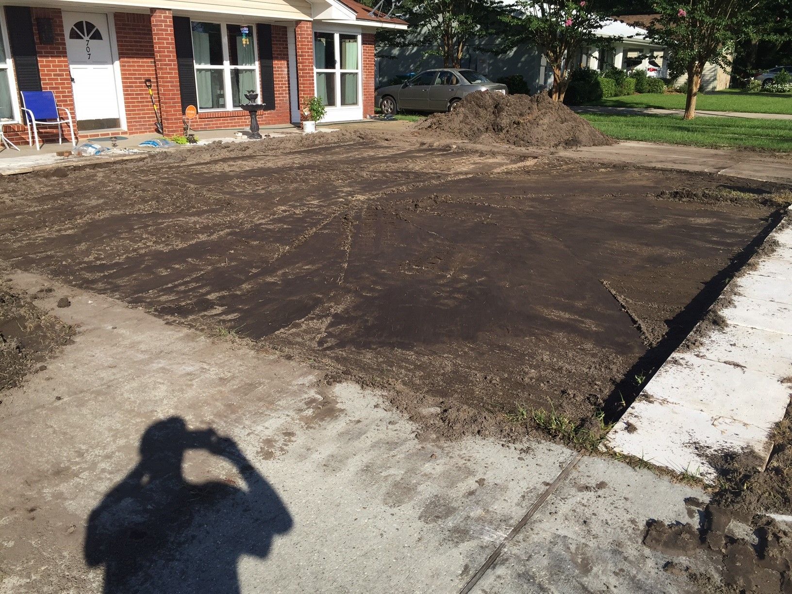 Freshly leveled dirt area beside a brick house, with a sidewalk and curb in the foreground