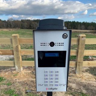 Outdoor keypad access terminal with screen and camera beside a wooden fence under a cloudy sky