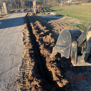 Backhoe digging a long trench beside a paved road in a grassy field