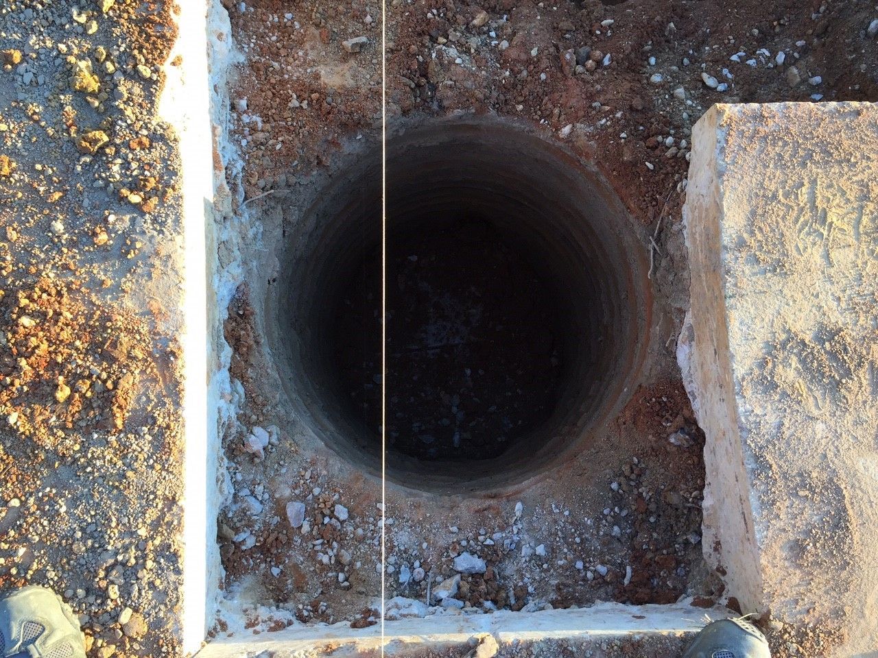 Top-down view of a dark circular hole in concrete beside rough stone edges and gravel.