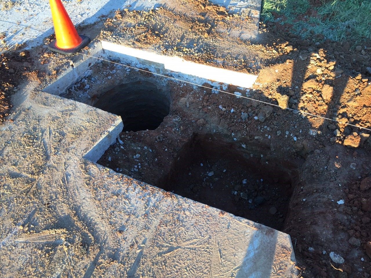Open utility trench or hole in dirt, with an orange cone nearby and wooden boards on the edge