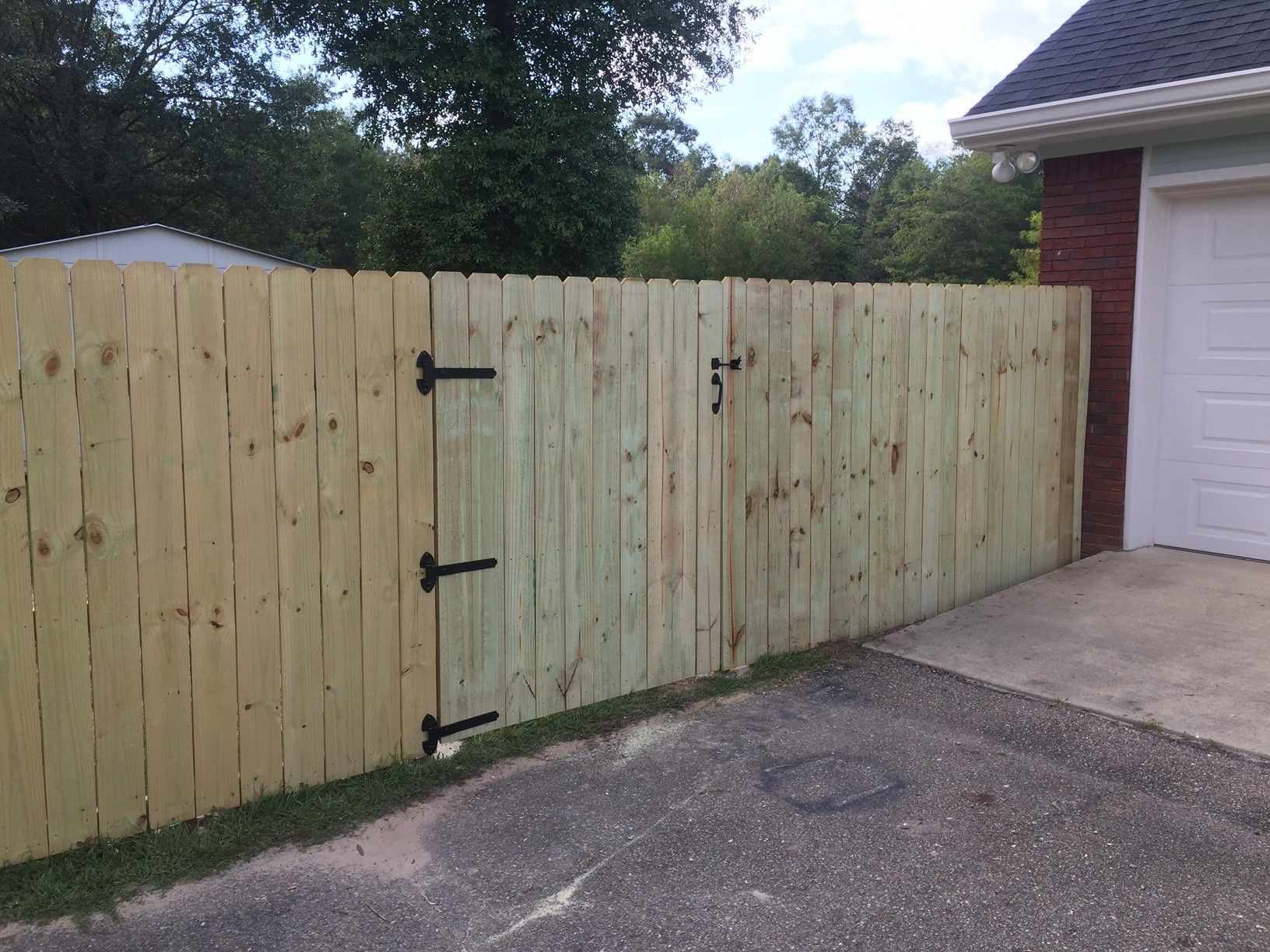 Wooden privacy fence beside a driveway, with black hinges and a red brick garage on the right.
