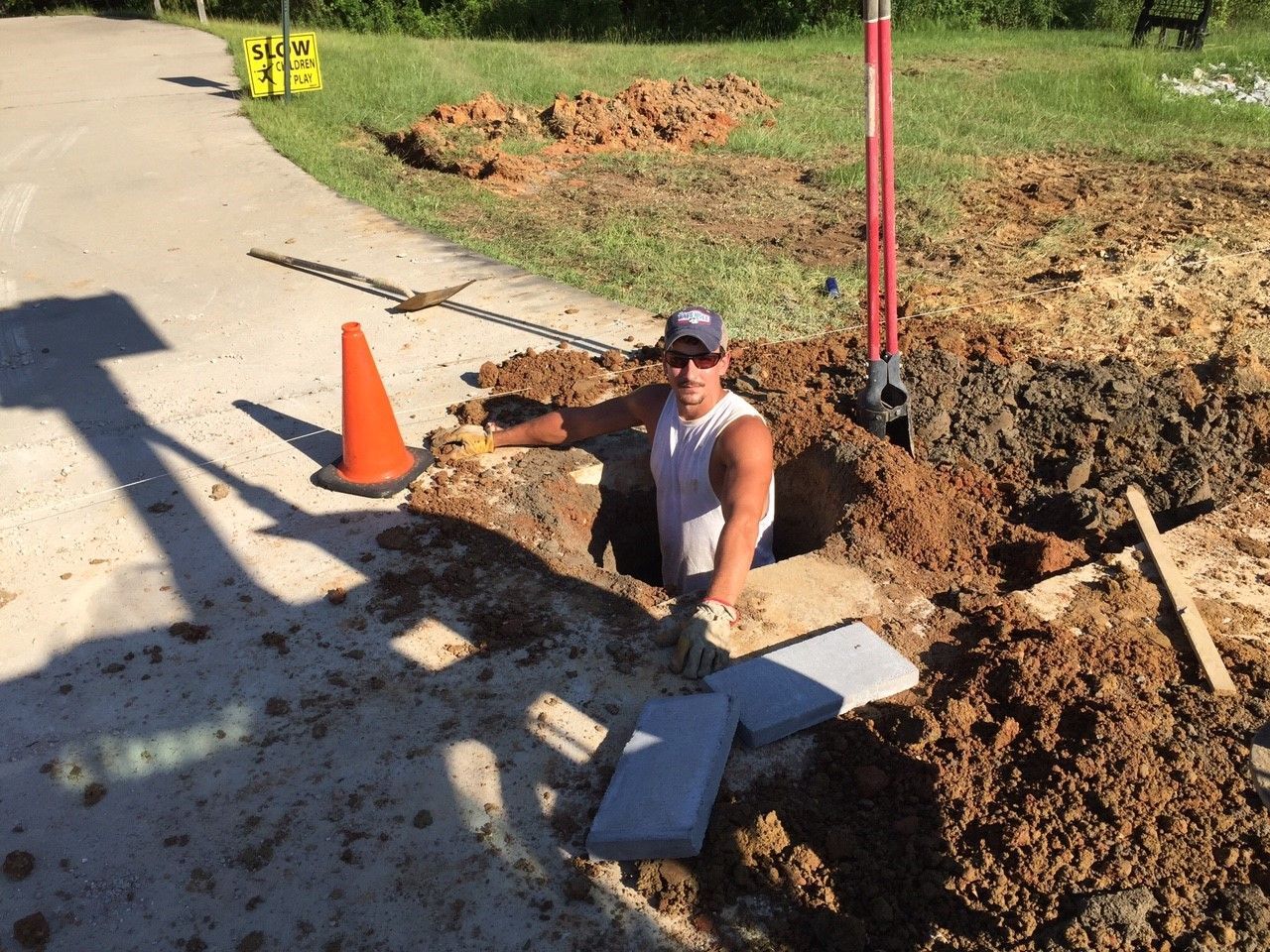 Worker in a trench beside a road repair site, with orange cone and dirt piles.