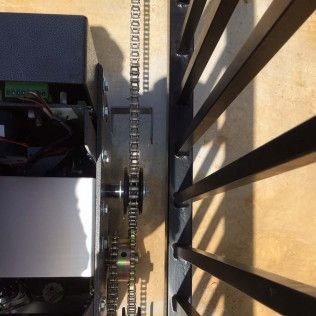 Close-up of a metal chain and gear mechanism beside black slats on a beige surface.