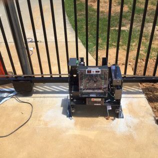 Small black device mounted on a concrete patio beside a metal gate, with a power cord attached