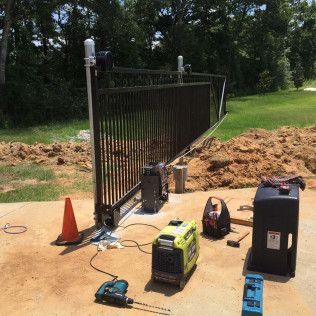 Black metal gate being installed outdoors with orange cone and power tools on a dirt driveway.