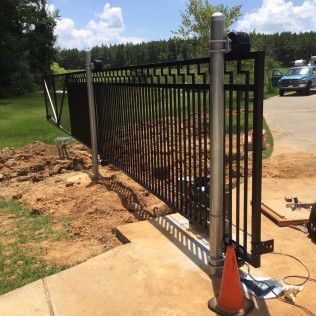 Black metal fence gate with posts at a driveway, partly installed beside a concrete walkway and dirt work area.