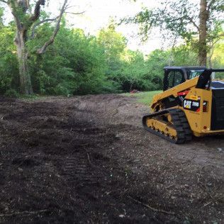 Yellow skid steer clearing dirt in a wooded area near a gravel path