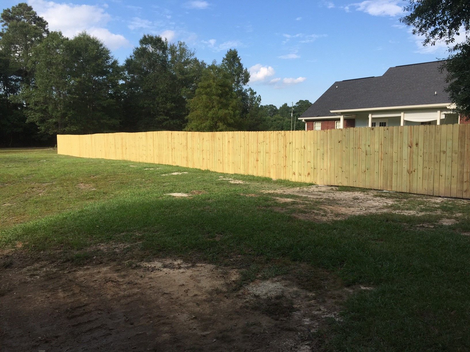 Long wooden fence beside a grassy yard with trees and a house under a blue sky