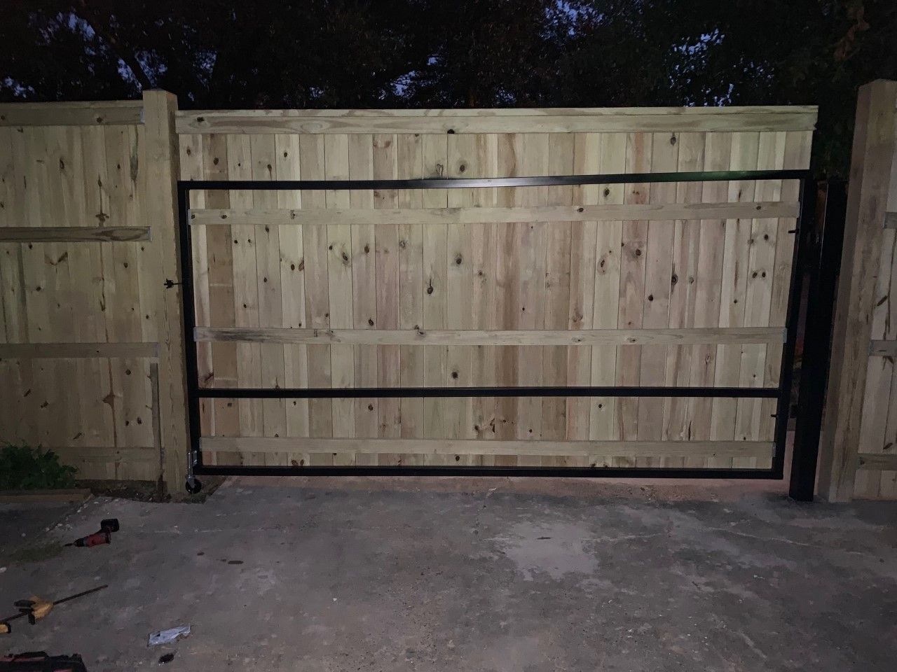 Wooden driveway gate at night, closed against a fenced yard.