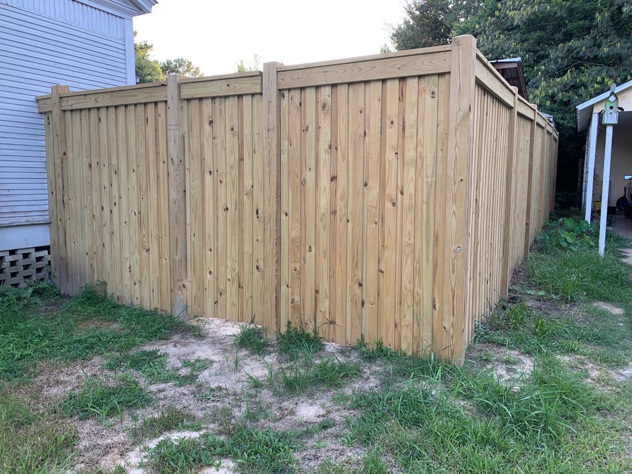 Wooden privacy fence enclosing a yard beside a house, with grass and dirt in front.