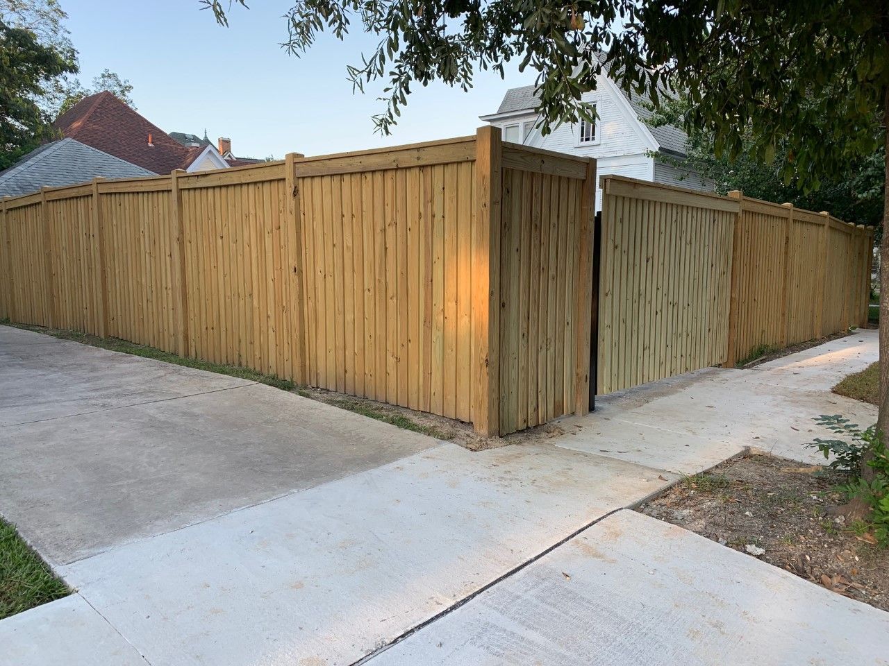 Wooden privacy fence along a corner sidewalk and driveway, with trees overhead and a house roof visible