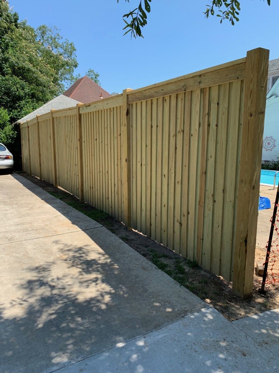 Long wooden privacy fence along a driveway in bright sunlight, with trees and a parked car nearby