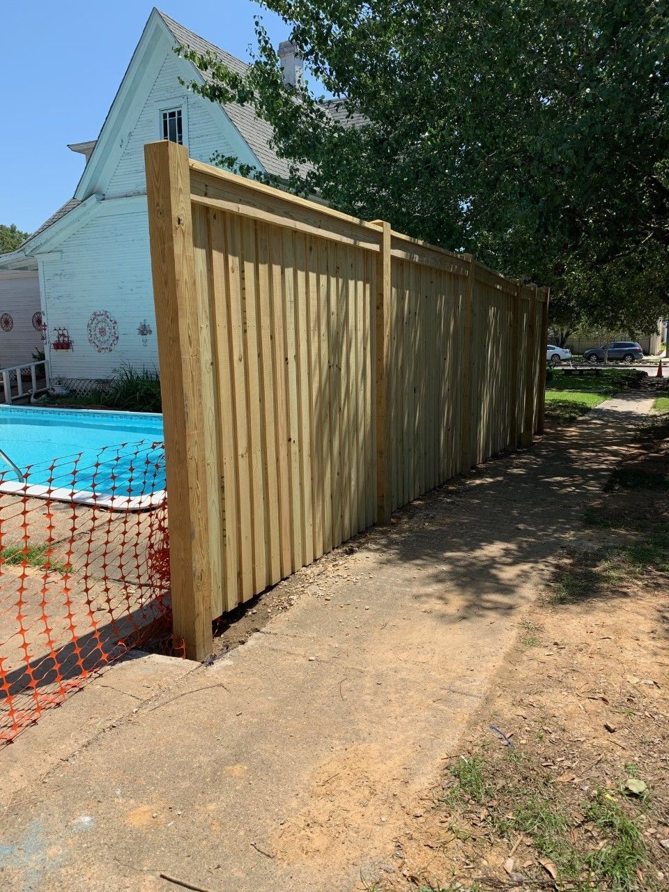 Wooden privacy fence beside a blue pool, with a dirt path and trees in a sunny yard.