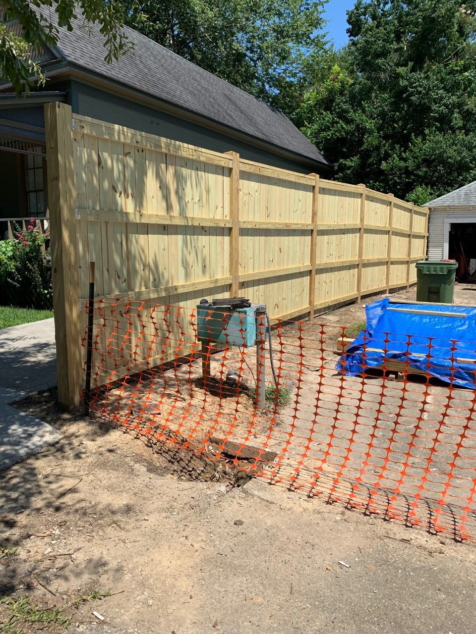 Wooden fence under construction beside a driveway, with orange safety netting and a blue tarp.