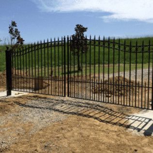 Black metal gate along a gravel drive, opening to a grassy hillside under a blue sky.