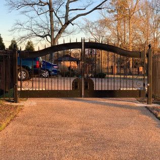 Closed black wrought-iron gate at driveway entrance with a blue pickup truck beyond it at sunset
