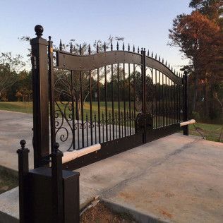 Black wrought-iron driveway gate with decorative arches at a paved entrance, beside a grassy yard.