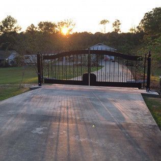 Closed black gate at the end of a driveway at sunset, with trees and a house in the background
