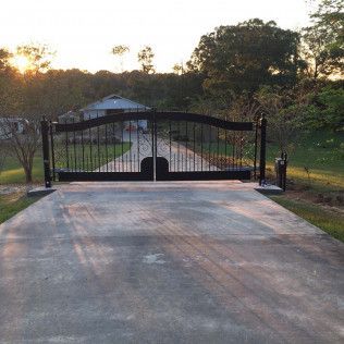 Driveway leading to a black metal gate at sunset, with trees and a house beyond.