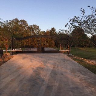 Concrete driveway leading to a closed black gate, bordered by trees at sunset.