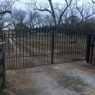 Black iron gate across a concrete driveway, with leafless trees and a fenced yard beyond.