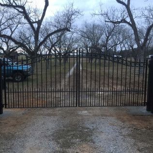 Closed black iron gate across a gravel driveway with bare trees and a blue car in the background