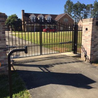 Black metal driveway gate beside brick pillars, opening to a house with a lawn and parked red car.