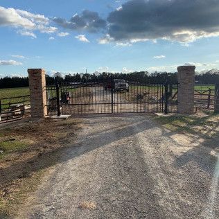Gated gravel driveway with brick pillars leading to a rural property under a partly cloudy sky