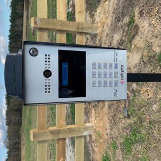 Outdoor silver intercom kiosk with keypad and screen beside a wooden fence on dirt ground