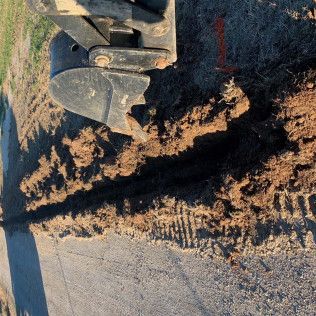 Excavator bucket cutting a dirt trench beside a paved road, leaving a rough brown edge.
