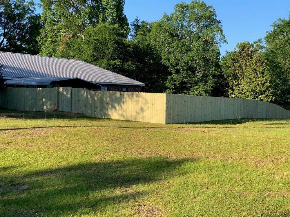 Grassy field beside a pale yellow fence and a metal-roofed building with trees in the background