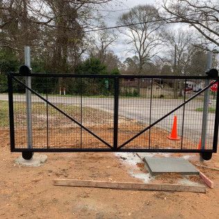 Black metal gate blocking a muddy driveway, with an orange cone and road beyond