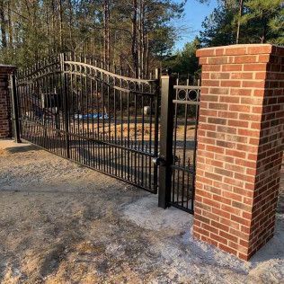 Black metal driveway gate beside a brick pillar, with trees and a gravel entrance behind it.