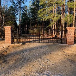 Closed black wrought-iron gate between brick pillars on a gravel driveway in a wooded area