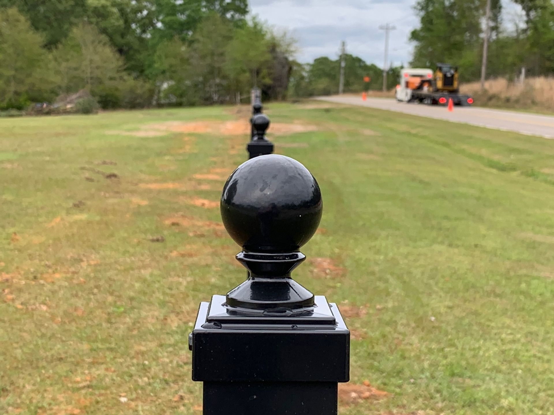 Black fence post in foreground of a grassy roadside with workers and machinery in the distance