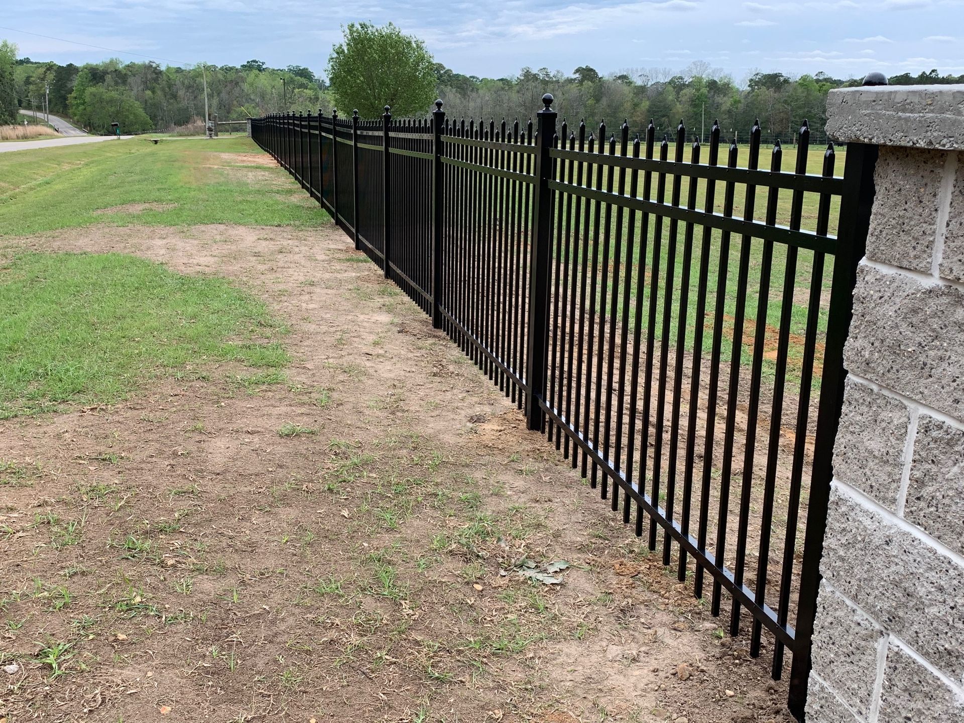 Black metal fence along a grassy dirt path beside a stone wall under a cloudy sky