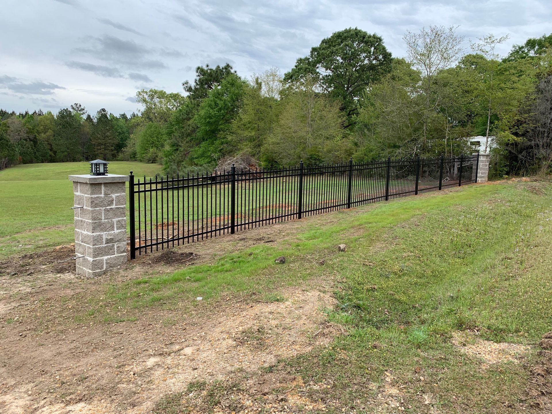 Black metal fence between stone pillars along a grassy roadside with trees and cloudy sky