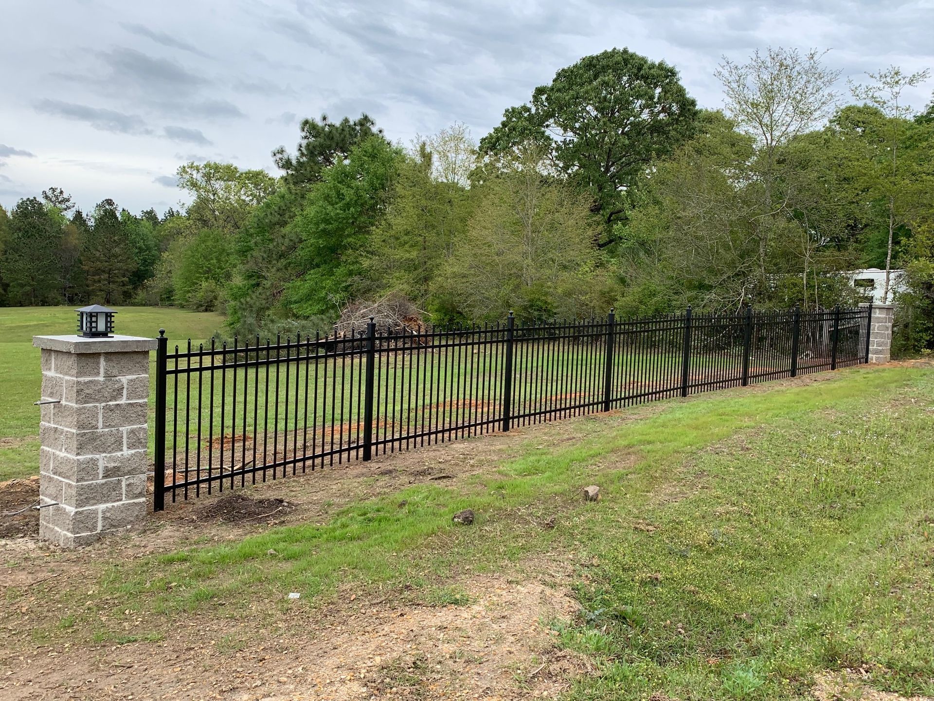 Black iron fence beside a grassy slope, with stone pillars and trees in the background.