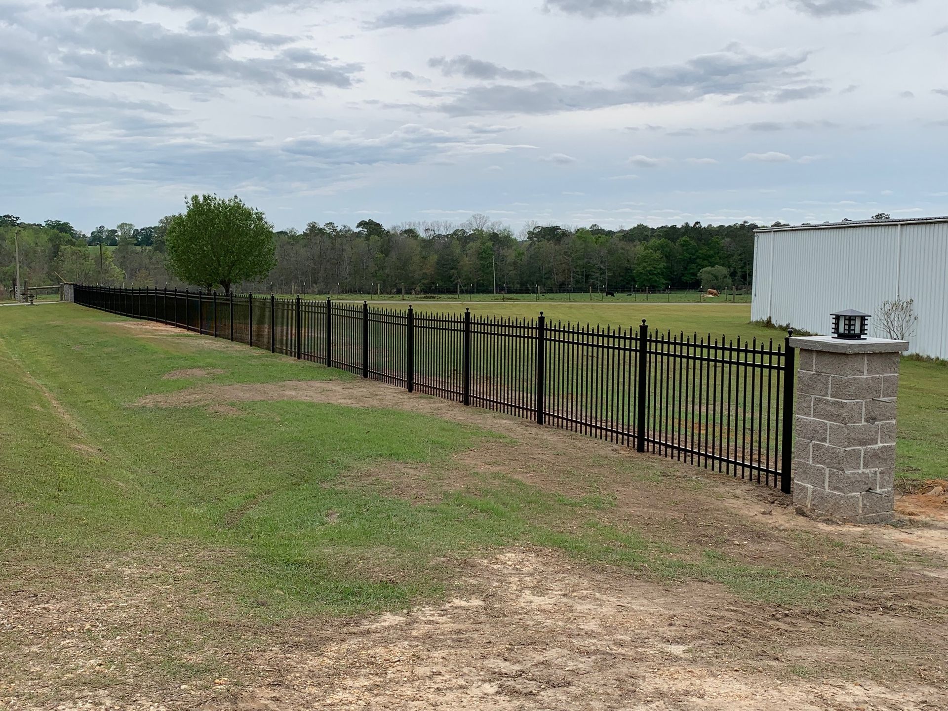 Black metal fence beside a grassy field and stone gatepost under a cloudy sky.