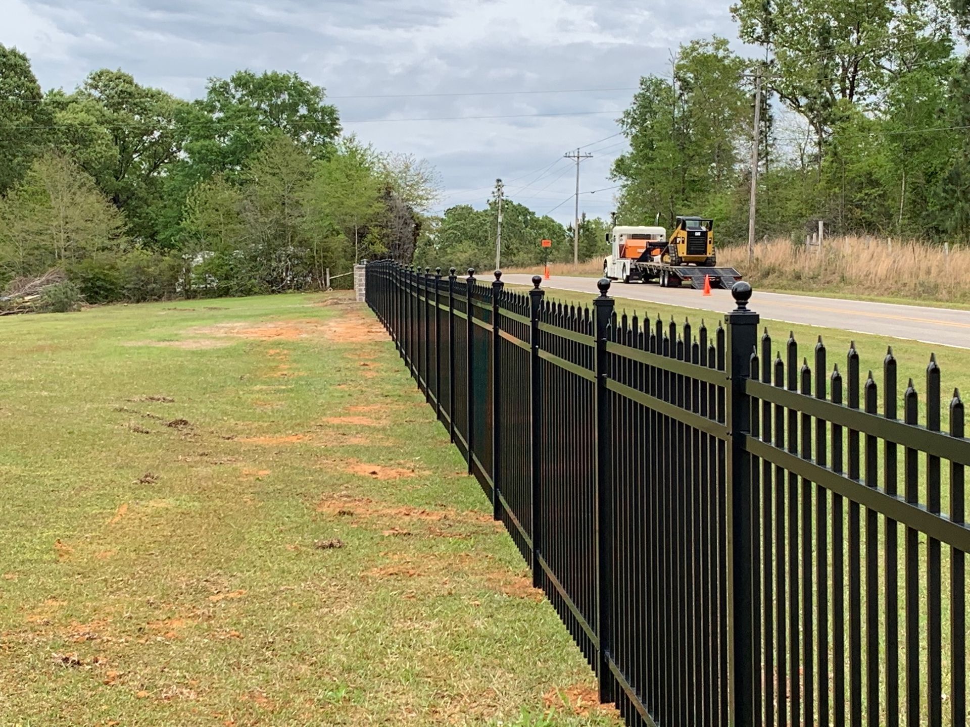 Black metal fence along a grassy roadside with workers and a utility truck in the distance