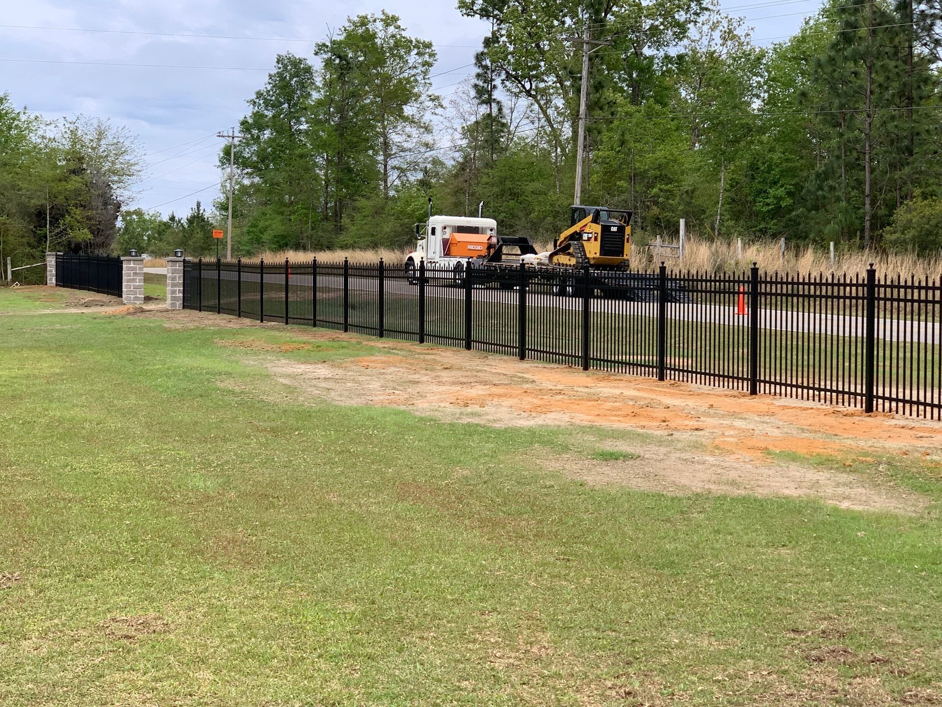 Black metal fence beside a grassy yard; a white utility truck and yellow excavator work among trees.
