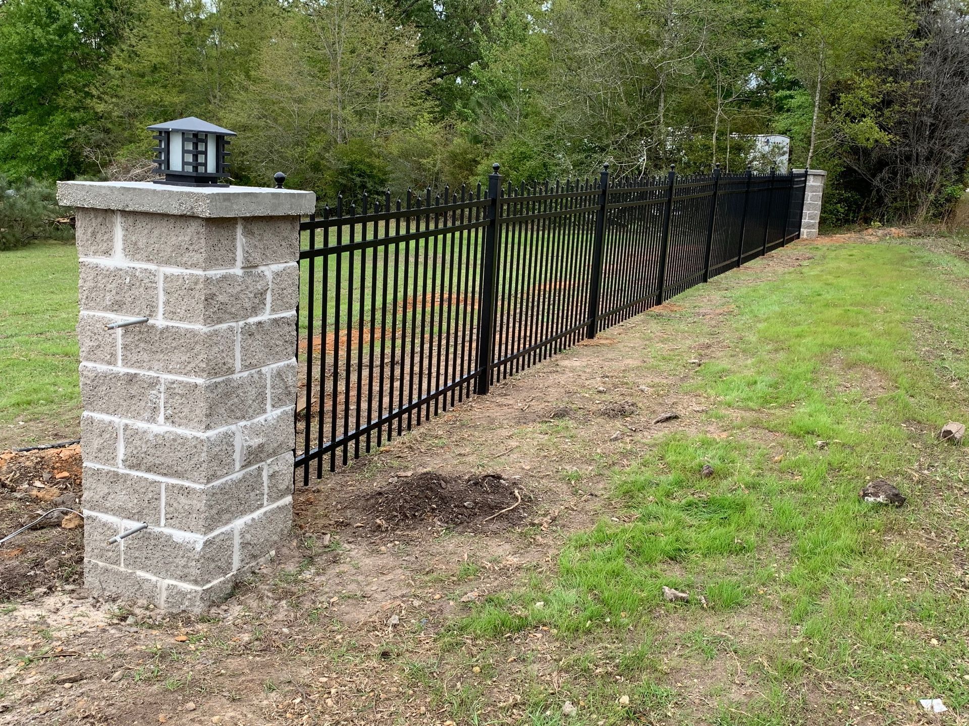 Black metal fence beside a stone pillar along a grassy slope and tree-lined yard