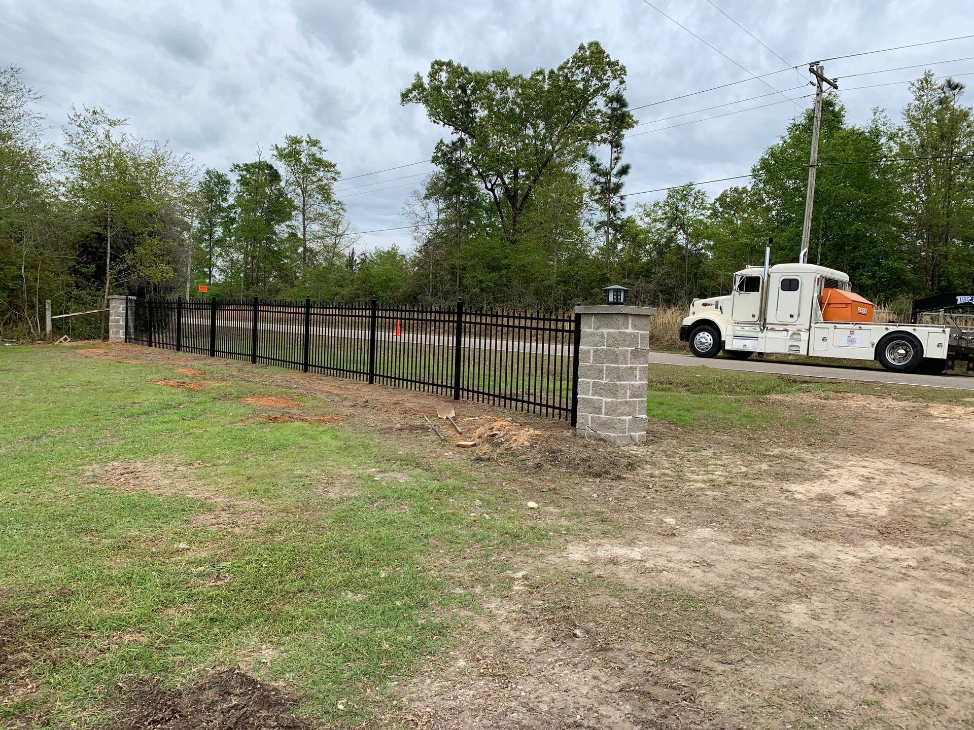 Black metal fence beside a dirt path, with a white truck parked near a stone gate pillar under cloudy skies