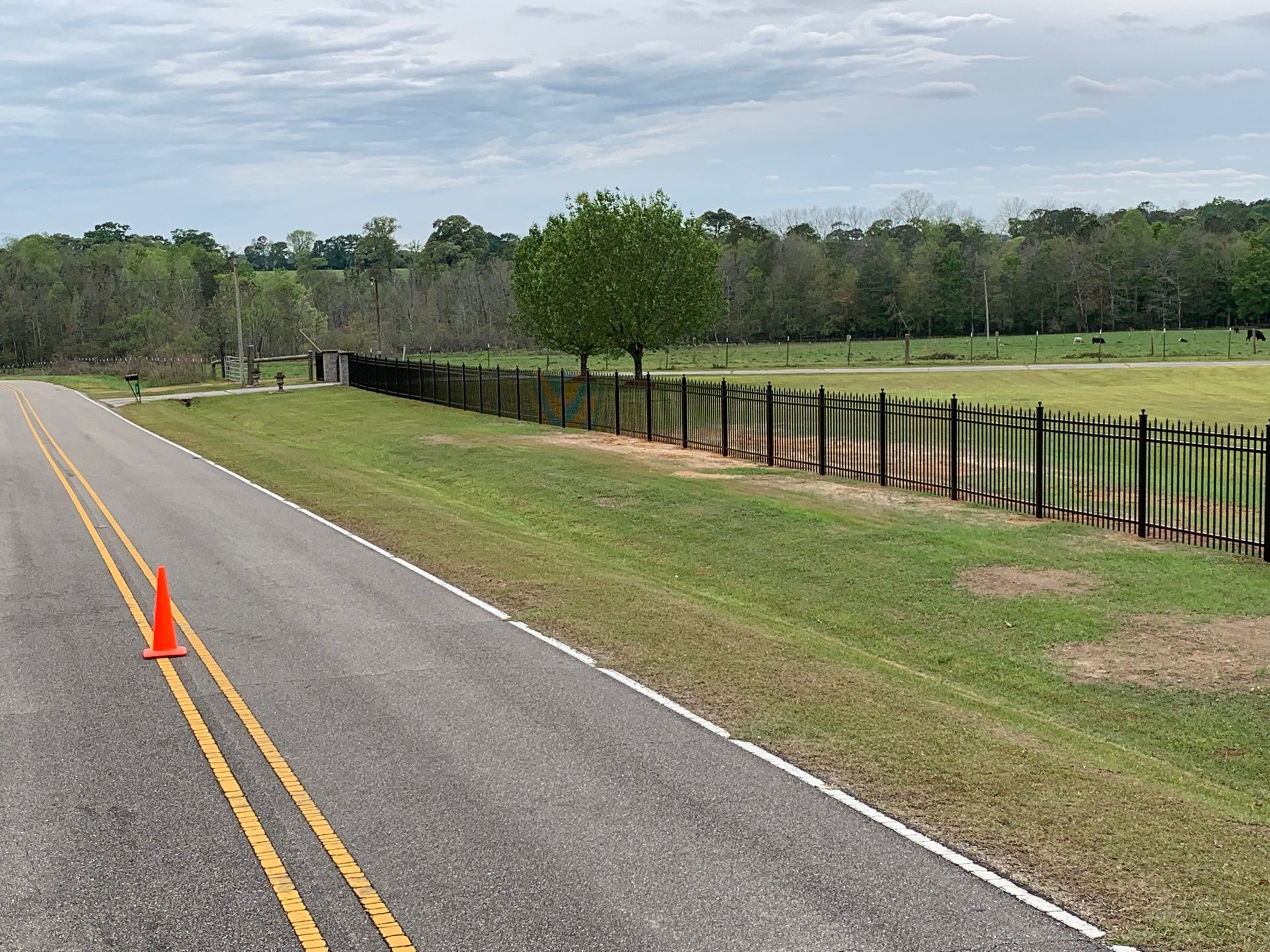 Empty road with orange cone beside a fenced green pasture on a cloudy day